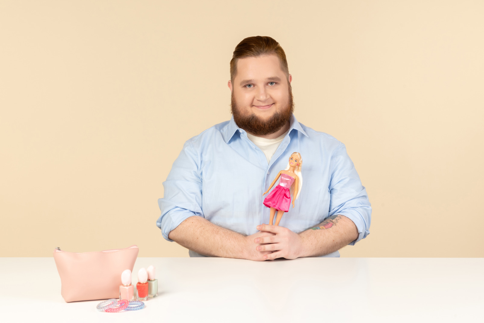 Smiling shy young big man sitting at the table and holding barbie doll