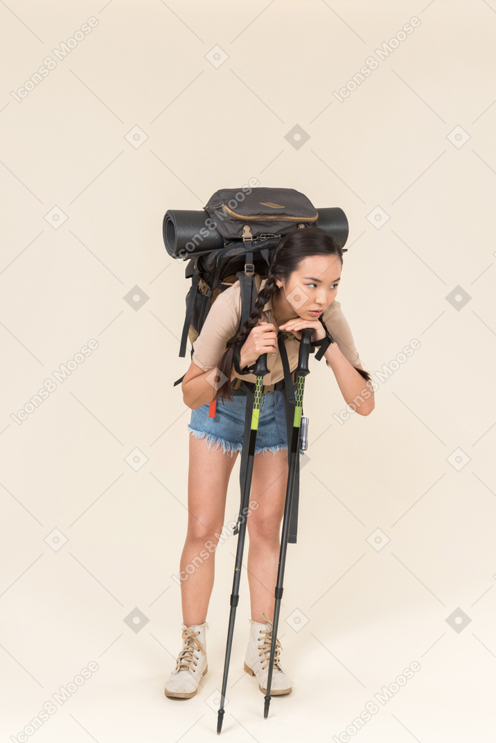 Stanco giovane escursionista donna in piedi con enorme zaino e appoggiandosi su bastoncini da trekking