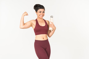 Joven mujer india en ropa deportiva sosteniendo la botella deportiva y mostrando los músculos