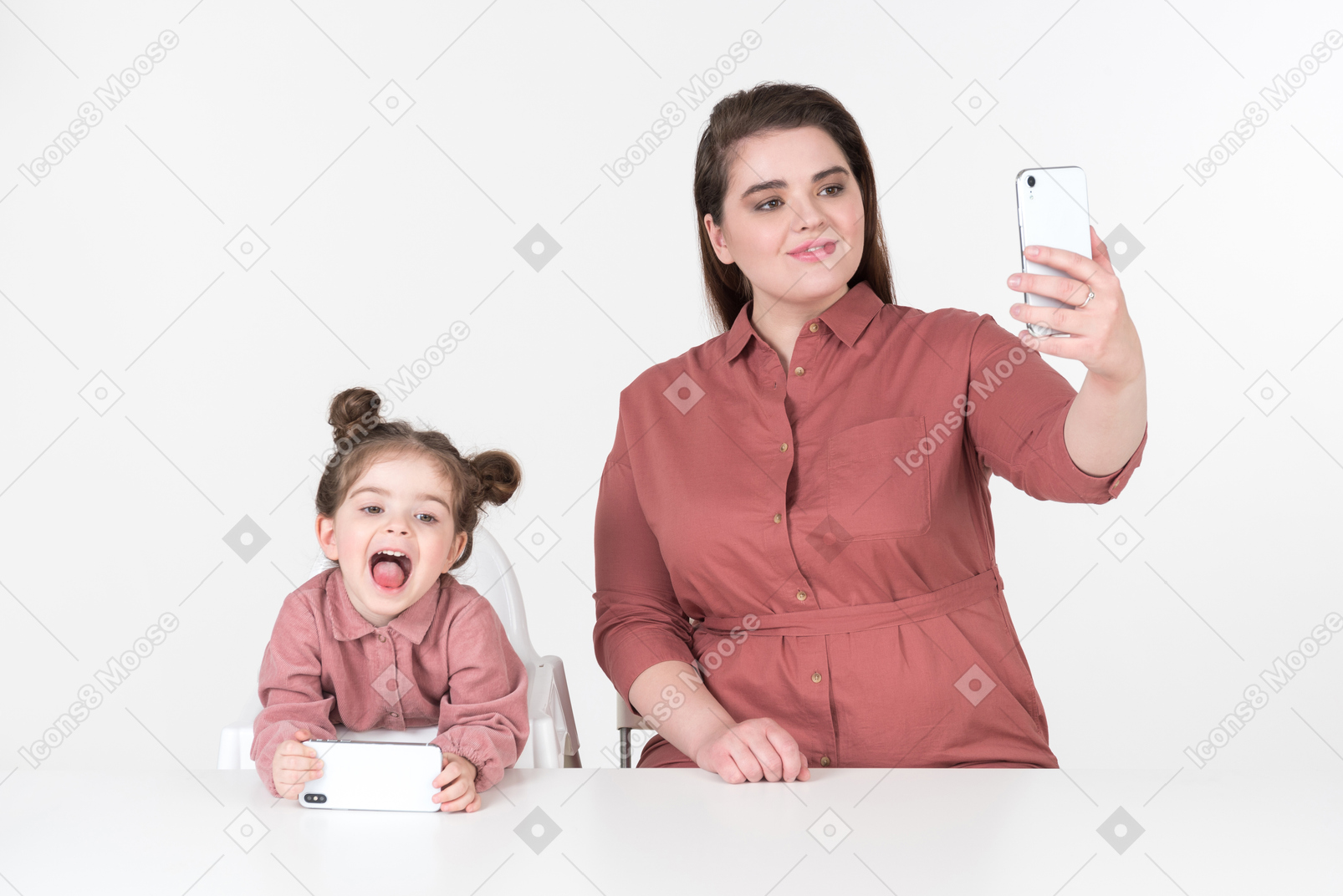 Mother and her little daughter, wearing red and pink clothes, sitting at the dinner table with smartphones in their hands