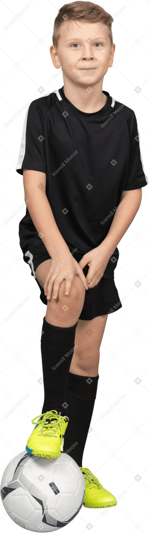 一个微笑的儿童男孩穿着足球制服，将他的脚放在球上，抚摸着膝盖的前视图