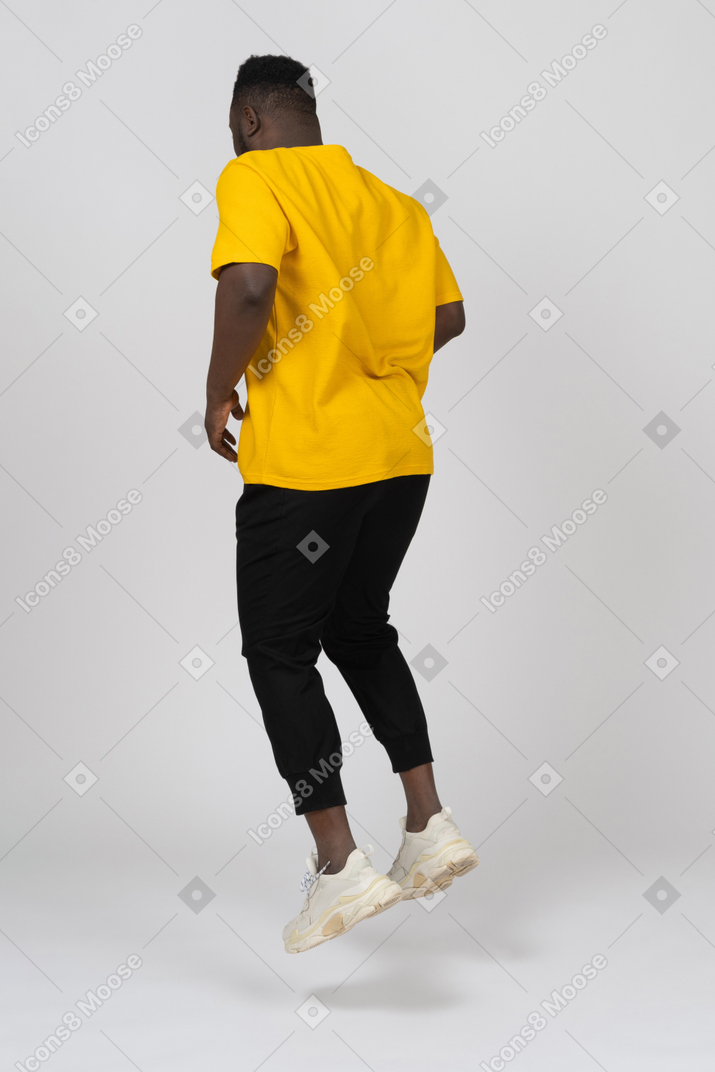 Vista posterior de tres cuartos de un joven de piel oscura saltando en camiseta amarilla