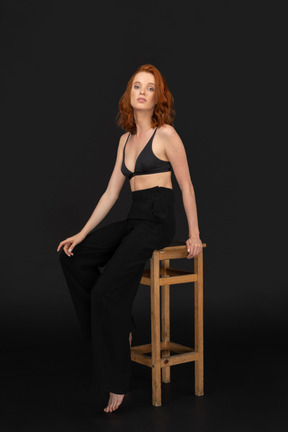 A side view of the beautiful woman dressed in black pants and bra, sitting on the wooden chair, holding her hand on the knee and looking to the camera