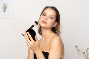 Side view of a young woman applying face powder while holding a mirror