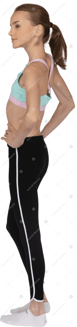 Dreiviertel-rückansicht eines jugendlichen mädchens in sportbekleidung, das hände auf hüften legt und stolz beiseite schaut