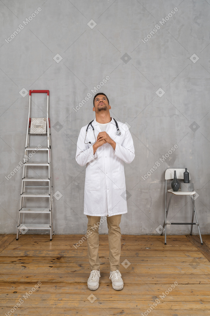 Vista frontal de um jovem médico orando em uma sala com escada e cadeira