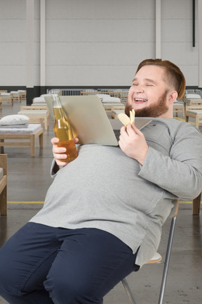 A man sitting in a chair holding a beer and a laptop