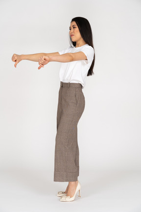 Vista di tre quarti di una giovane donna in calzoni e maglietta che mostra i pollici verso il basso