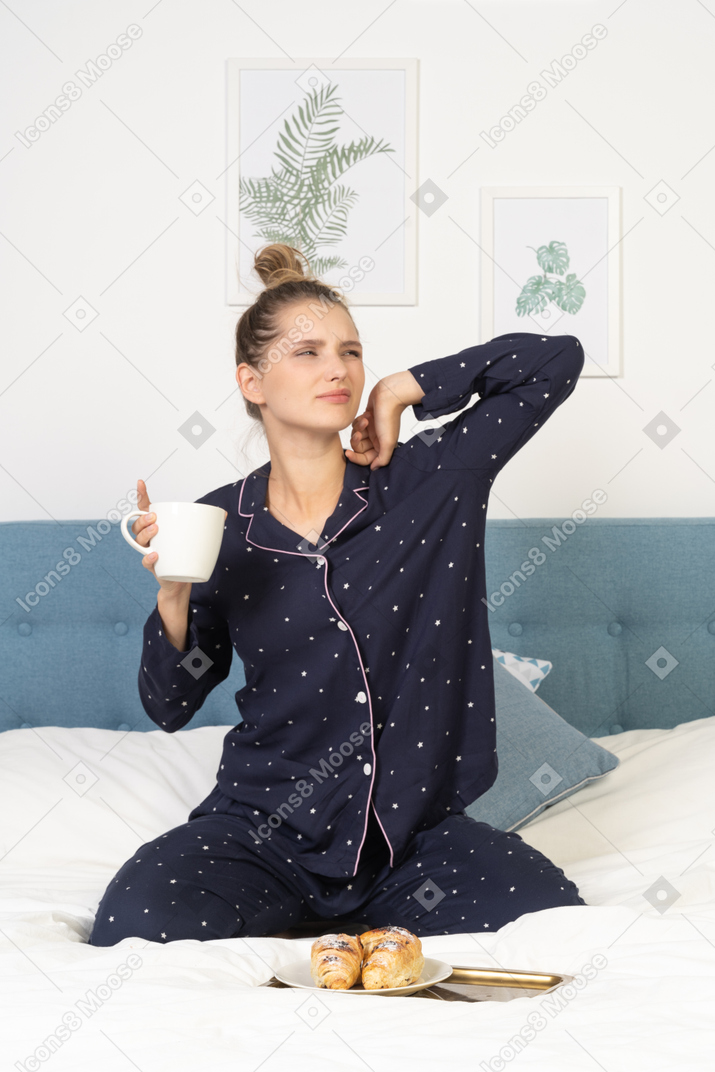 Front view of a stretching young lady in pajamas holding a cup of coffee while sitting in bed