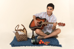 Junger kaukasischer kerl, der nahe picknickkorb auf der decke sitzt und gitarre spielt