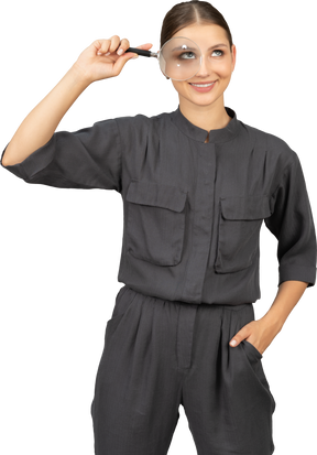 Front view of a smiling young woman in a jumpsuit holding a magnifying glass