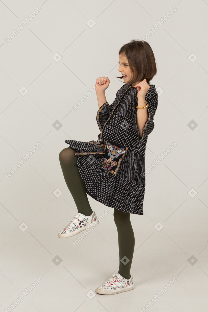 Side view of a happy little girl in dress clenching fists and raising leg