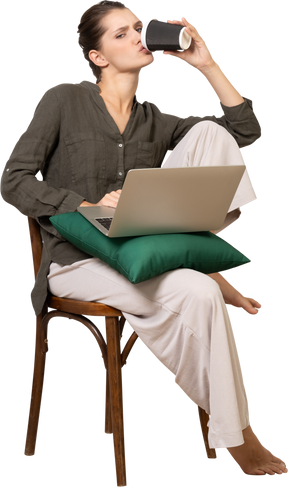 Vista frontale di una giovane donna seduta su una sedia che tiene in mano il suo laptop e beve caffè