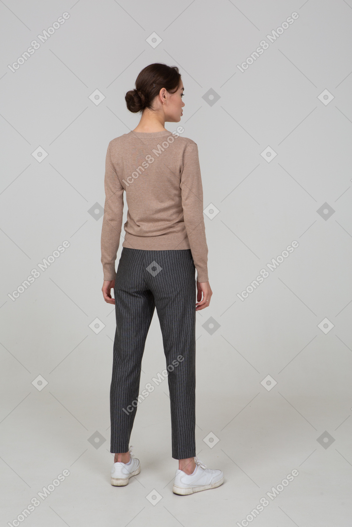 Three-quarter back view of a young lady standing still in pullover and pants
