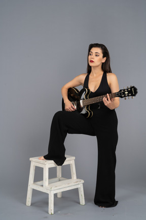 Vue de trois quarts d'une jeune femme en costume noir tenant la guitare et mettant la jambe sur le tabouret