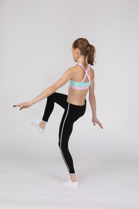 Три четверти сзади девушки-подростка в спортивной одежде, протягивающей руки и поднимающей ногу