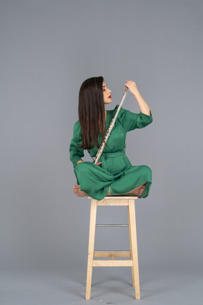 Девушка в полный рост смотрит на кларнет, сидя со скрещенными ногами на деревянном стуле