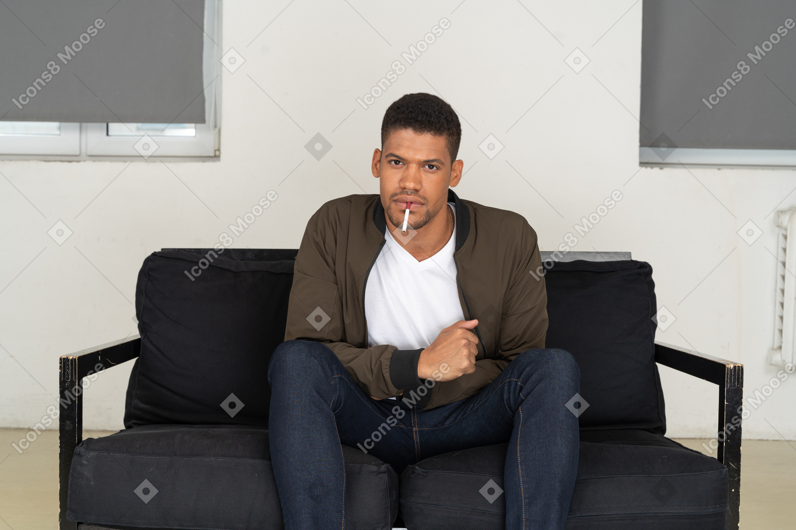 Vista frontal del joven sentado en un sofá y sosteniendo el cigarrillo en la boca