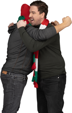 Вид сбоку двух эмоционально обнимающихся футбольных фанатов мужского пола, сжимающих кулаки