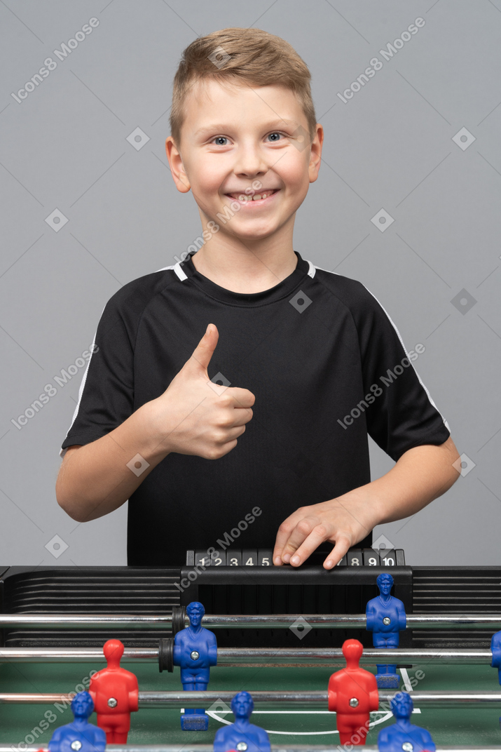 Vista frontal de um menino jogando pebolim e mostrando o polegar