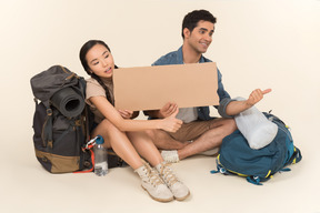 Young interracial couple of hitchhikers holding paper card and showing hitchhiking gestures