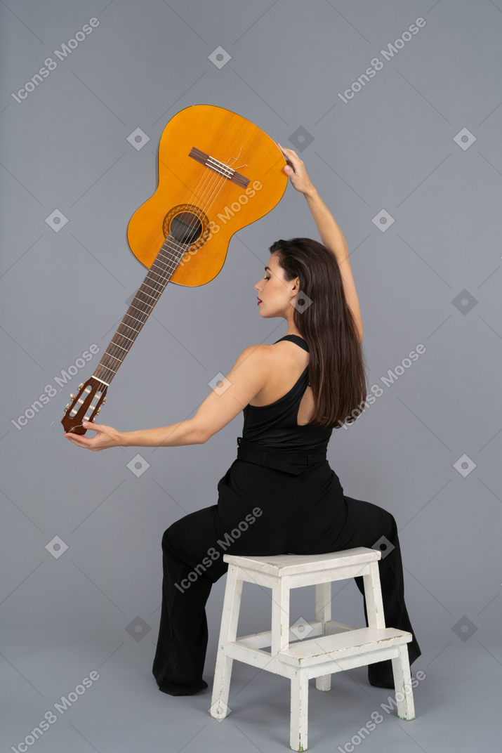 Young woman holding a guitar high while sitting on a stool