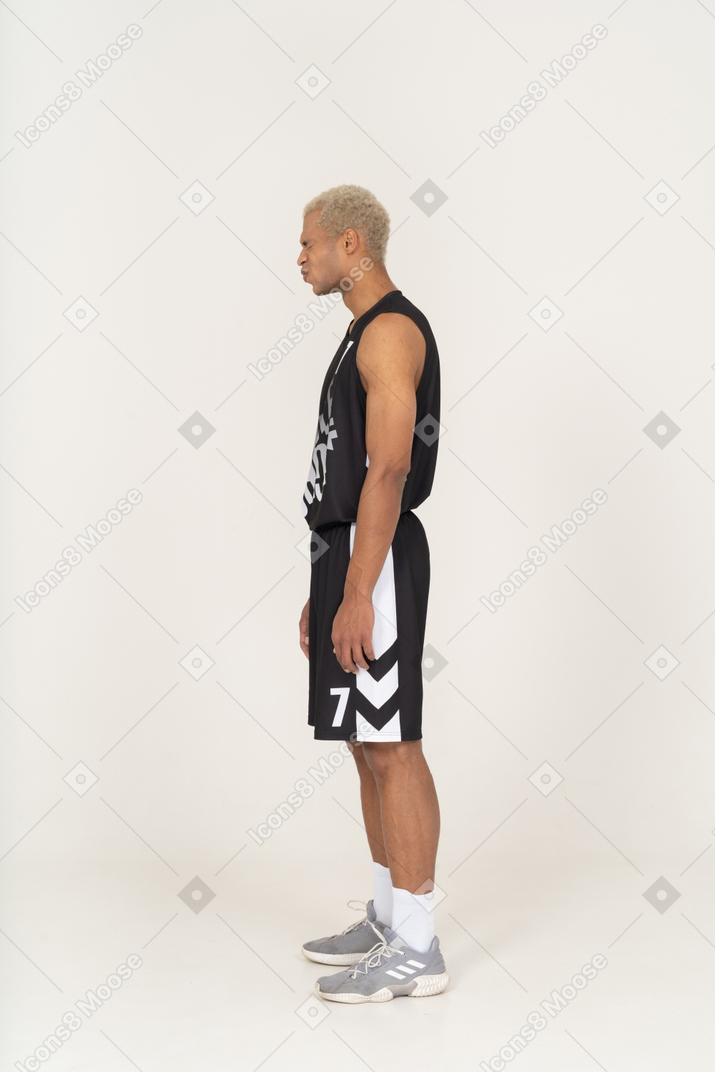 Vista lateral de un joven jugador de baloncesto masculino disgustado de pie y presionando los labios