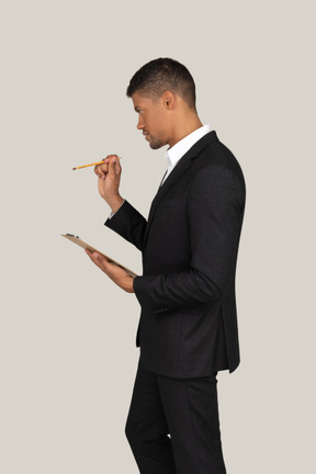 Standing in profile young man in black suit holding folder and pencil