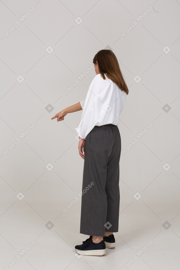 Вид сзади три четверти молодой леди в офисной одежде указывая пальцем