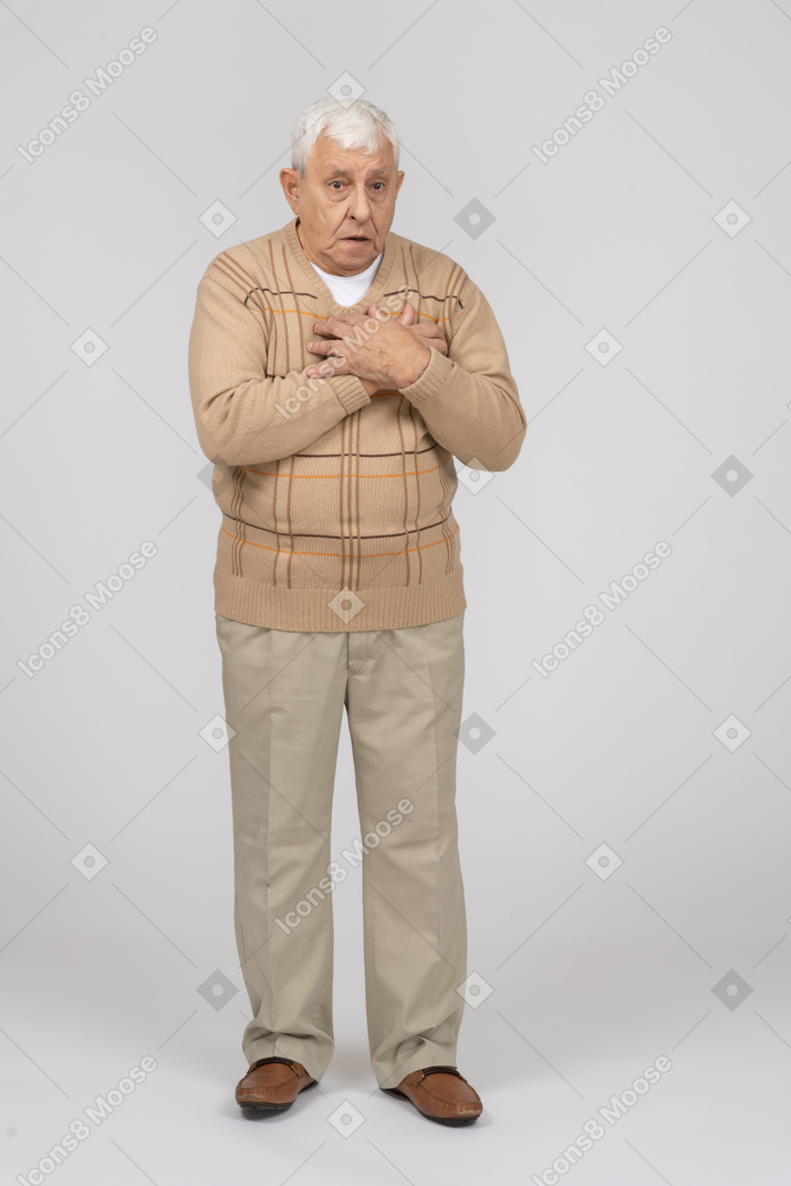 Front view of an impressed old man in casual clothes standing with hands on chest and looking at camera