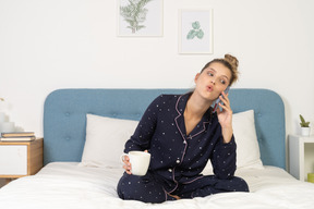 Na frente de uma jovem mulher de pijama, sentada na cama, segurando a xícara e fazendo uma ligação