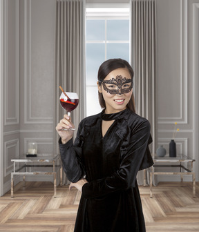 Красивая азиатская женщина в черном бархатном платье и маскарадной маске стоит в современной комнате с бокалом коктейля в руке