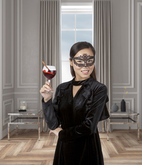 Beautiful asian woman in a black velvet dress and a masquerade mask standing in a modern-looking room with a glass of cocktail in her hand