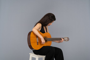 Vue de trois quarts d'une jeune femme assise en costume noir à jouer de la guitare et regardant vers le bas