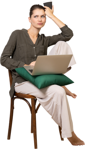 Front view of a confused young woman wearing home clothes sitting on a chair with a laptop and coffee