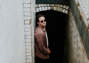 Side view of a young man in black sunglasses standing near the wall and looking at camera