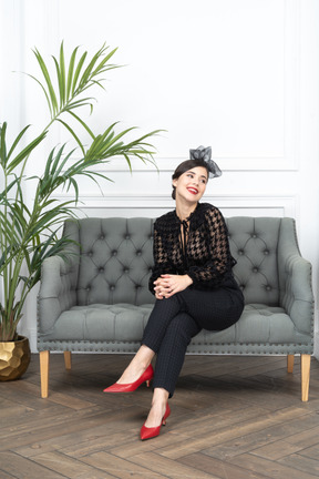 A smiling woman in black outfit sitting on a couch next to a potted plant 