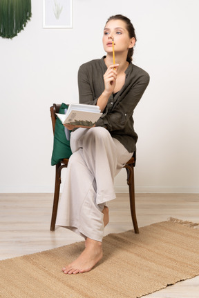 Front view of a thoughtful young woman wearing home clothes sitting on a chair with pencil and notebook