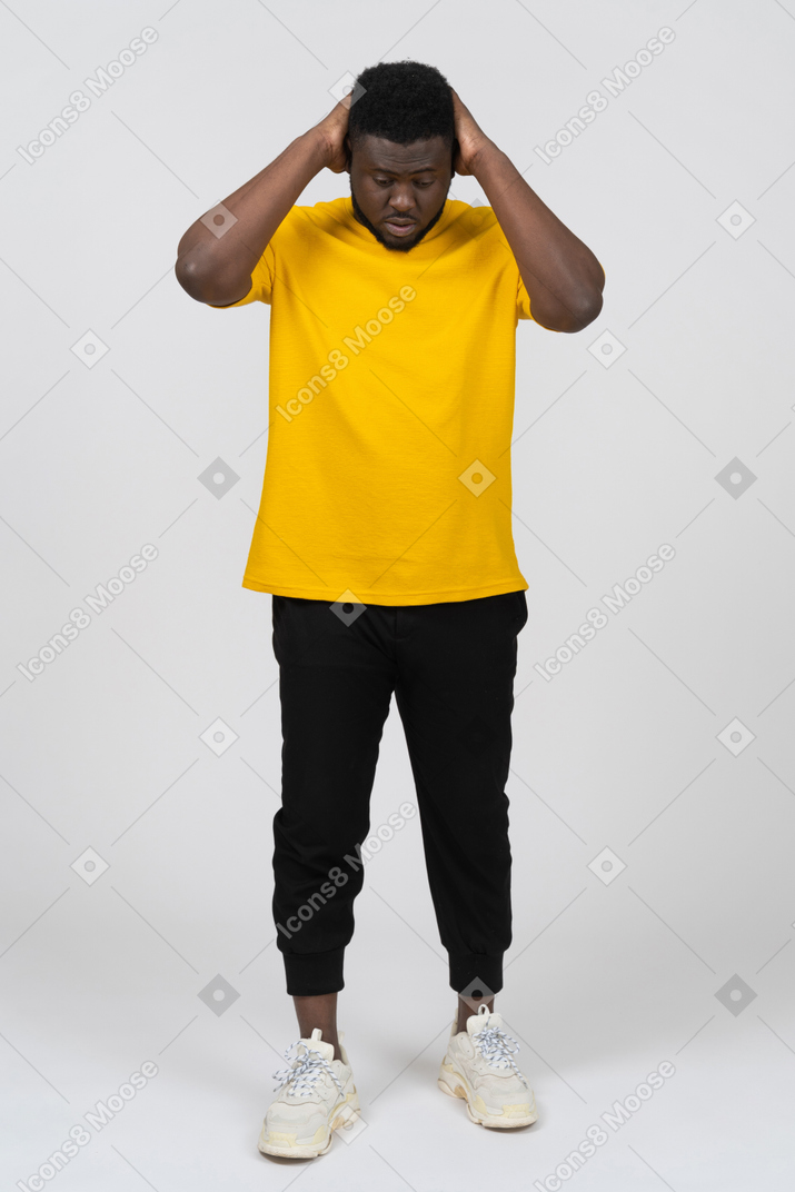 Front view of a young dark-skinned man in yellow t-shirt touching head & looking down