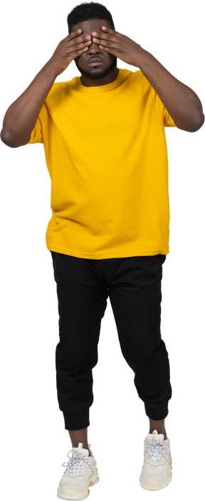 Front view of a young dark-skinned man in yellow t-shirt hiding his eyes