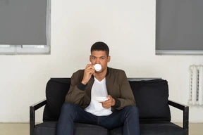 Vista frontal de un joven soñando sentado en un sofá mientras bebe café