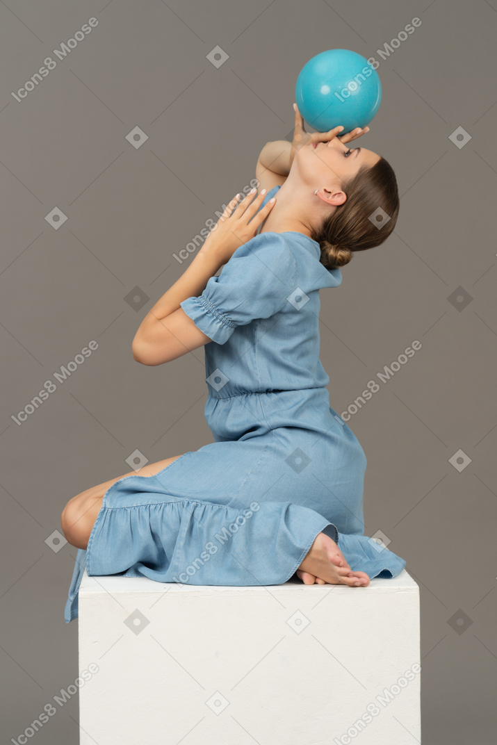 Vista lateral de una joven sentada en un cubo con una pelota en la cabeza