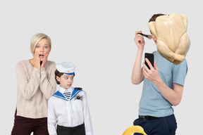 Shocked woman and boy looking at man with chicken head applying his makeup 