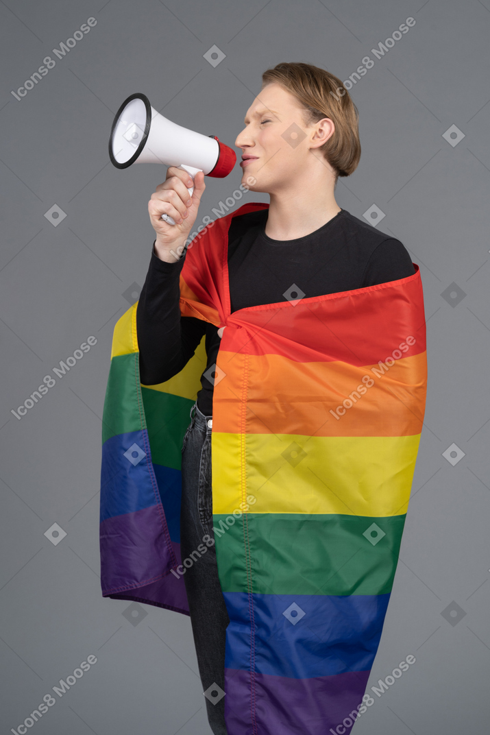 Pessoa não binária envolta em uma bandeira de arco-íris falando em um megafone