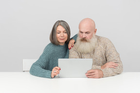 Aged couple sitting at table and looking at digital tablet