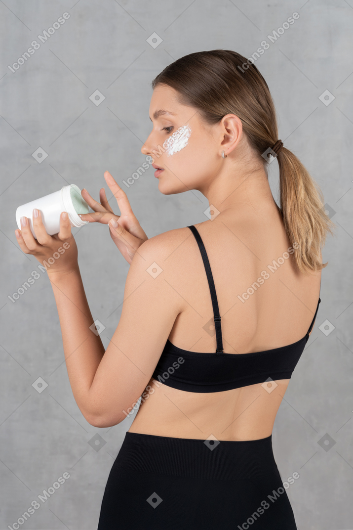 Vista traseira de uma jovem aplicando creme facial nas maçãs do rosto