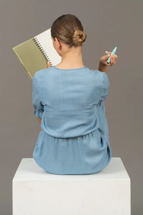 Back view of young woman in blue dress sitting on a cube and making notes