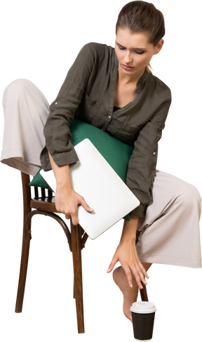 Vista frontal de uma jovem confusa, sentada em uma cadeira, segurando seu laptop e tocando a xícara de café