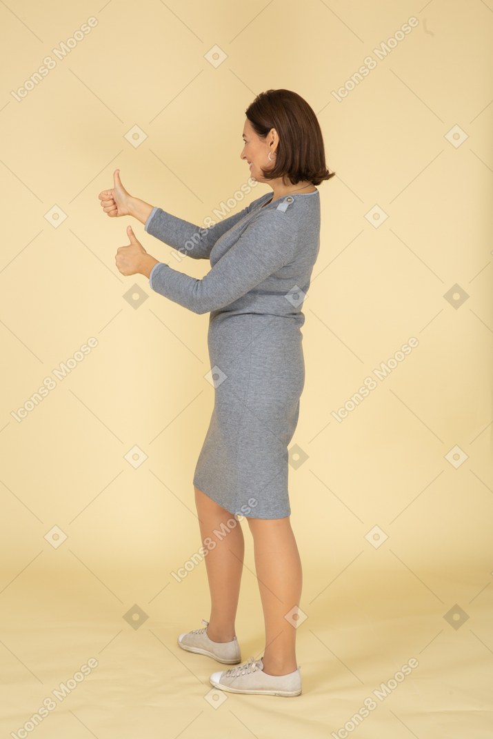 Femme en robe grise debout de profil et montrant les pouces vers le haut