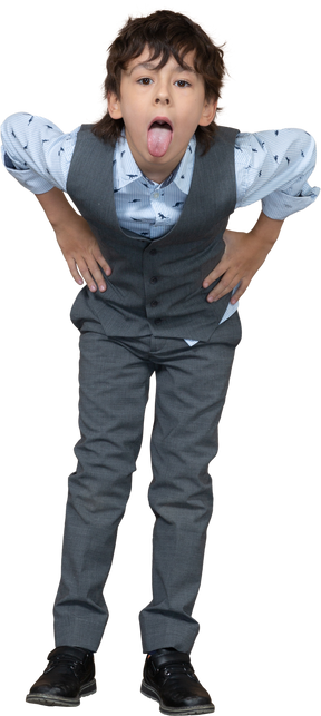 Vista frontal de um menino de terno cinza posando com as mãos nos quadris e mostrando a língua